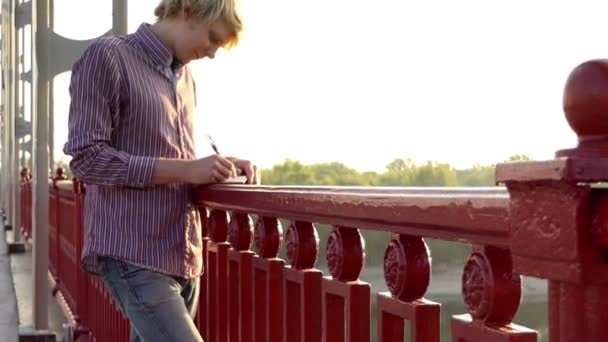 Junger Mann entwickelt Ideen und legt sie in einem Notizblock auf einer Brücke ab — Stockvideo