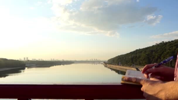 Blonder Mann steht und legt seine Ideen bei Sonnenuntergang auf einer Flussbrücke nieder — Stockvideo