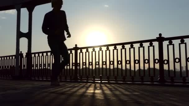 Молодой человек ходит и танцует на современном мосту на закате летом — стоковое видео