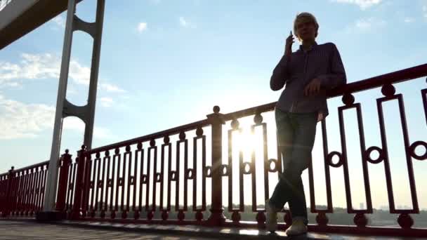 Jovem loiro goza de esplêndido pôr do sol em uma ponte do rio com um celular em Slo-Mo — Vídeo de Stock