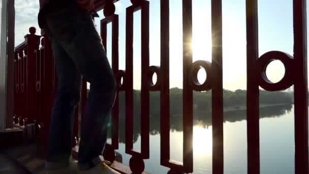 Блондинка людина стоїть на мосту і дістає свій мобільний на заході сонця в Slo-Mo — стокове відео