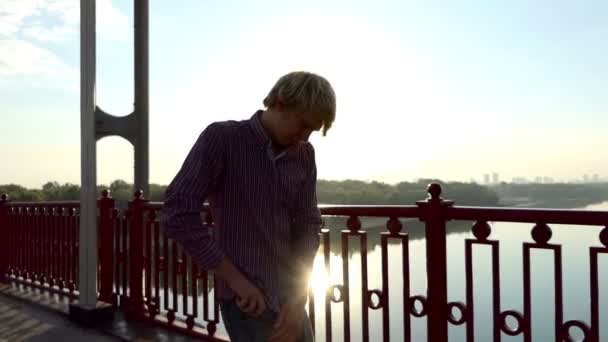 Young Man Walks Along the Bridge and Pulls Out His Mobile at Sunset in Slo-Mo — Stock Video