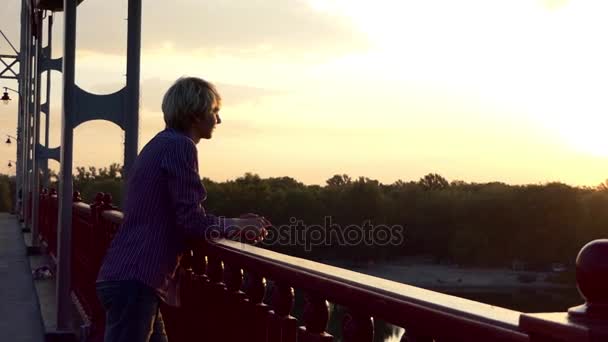 Junger Mann steht an einem sonnigen Tag auf einer Brücke über den Fluss Dnipro — Stockvideo