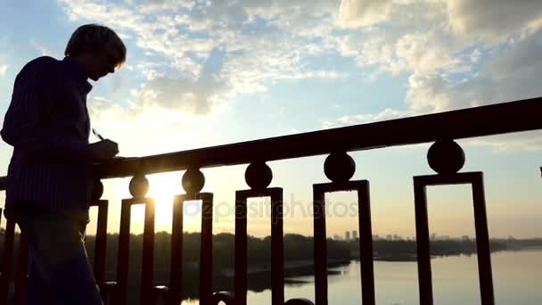 Romantische Mann steht und schreibt seine Ideen auf einer Flussbrücke bei Sonnenuntergang in slo-mo — Stockvideo