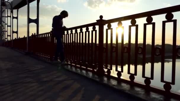 Joven se para y escribe sus ideas en un puente fluvial al atardecer — Vídeo de stock