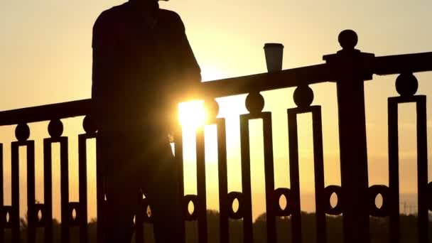 Красивий чоловік п'є каву з паперових склянок і користується захід сонця на мосту — стокове відео