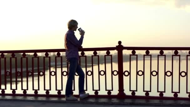 Hombre romántico bebe café de una taza de papel y disfruta de una puesta de sol en un puente en Slo-Mo — Vídeo de stock