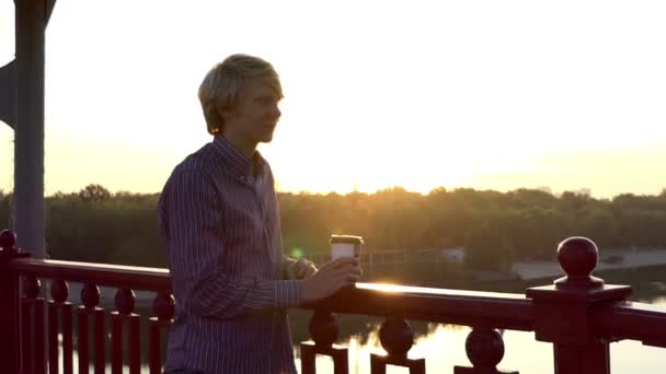 Happy Man boit du café dans une tasse en papier et profite d'un coucher de soleil sur un pont à Slo-Mo — Video