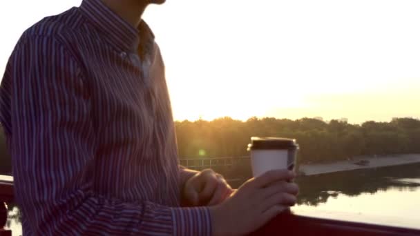 若い男は紙コップでコーヒーを飲みながら立ち橋に沈む夕日を楽しんでいます — ストック動画