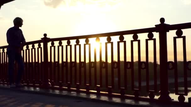 Jovem loiro olha para um esplêndido pôr do sol em uma ponte do rio com um celular em Slo-Mo — Vídeo de Stock