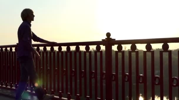 Jeune homme blond regarde un magnifique coucher de soleil sur un pont de rivière sur le Dnipro — Video