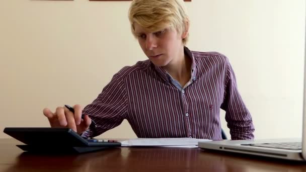 Блондин сочиняет некоторые документы и смотрит на свой калькулятор в офисе — стоковое видео