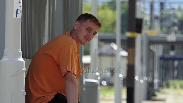 Un giovane uomo si siede su una stazione ferroviaria in estate. Fa l'occhiolino al rallentatore. — Video Stock