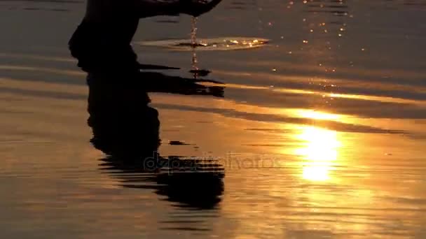 Homem jovem levanta água do rio em palmas e olha para ele em um pôr do sol dourado — Vídeo de Stock