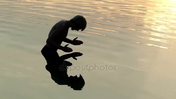 El hombre mira su reflejo, eleva el agua del río y la deja caer al atardecer — Vídeos de Stock