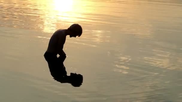 Человек смотрит на свое отражение, поднимает речную воду, и позволяет ей падать на закате — стоковое видео