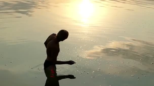 Joven se para en el agua del río y lanza una piedra plana al atardecer — Vídeos de Stock