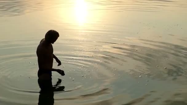 Rolig människa står i flodvatten och kastar en platt sten vid solnedgången i Slo-Mo — Stockvideo