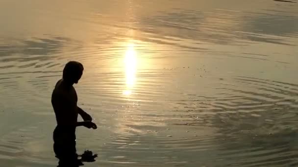 Slimme Man staat in rivierwater en gooit een platte steen bij zonsondergang — Stockvideo