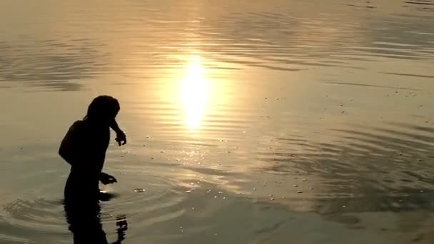 Szczęśliwy człowiek stoi w wodzie rzeki i zgłasza płaski kamień o zachodzie słońca w Slo-Mo — Wideo stockowe