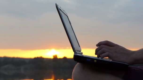 Mann tippt Finger auf einer PC-Tastatur im Freien auf einem Flussufer bei Sonnenuntergang — Stockvideo