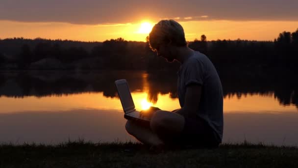 Kreativer Wan tippt auf seinem Laptop an einem Flussufer bei herrlichem Sonnenuntergang — Stockvideo