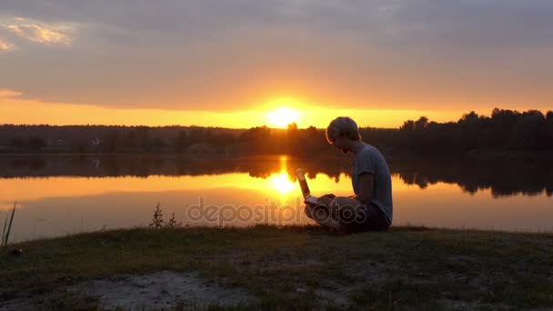 Talented Man Types on His Laptop on a River Bank at a Splendid Sunset — Stock Video