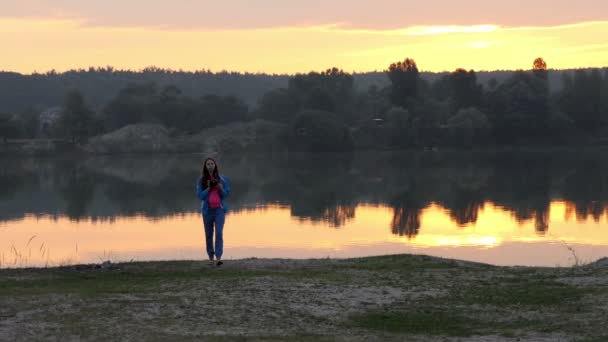 Těhotná žena chodí na břehu jezera, se dívá na její mobil, při západu slunce v Slo-Mo — Stock video