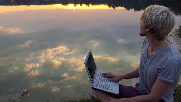 Творческий человек сидит со своей верхушкой на берегу реки на роскошном закате в Сло-Мо — стоковое видео