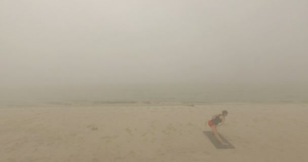 Mujer del yoga se para en una estera y dobla sus piernas en una playa de arena — Vídeo de stock