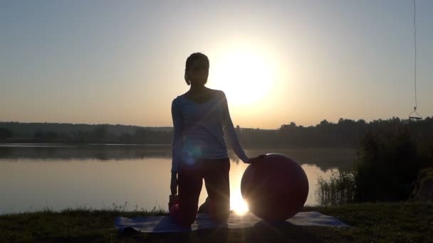 Una joven está de pie con una pelota de fitball en una orilla del lago — Vídeo de stock