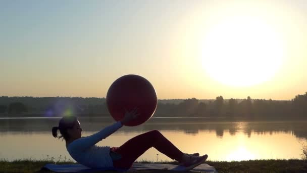 Een slanke vrouw ligt op een mat met een fitball in handen bij zonsondergang — Stockvideo