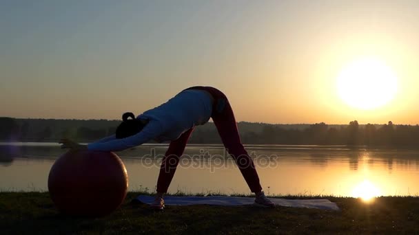 Uma mulher magra se levanta e rola seu fitball ao pôr do sol — Vídeo de Stock