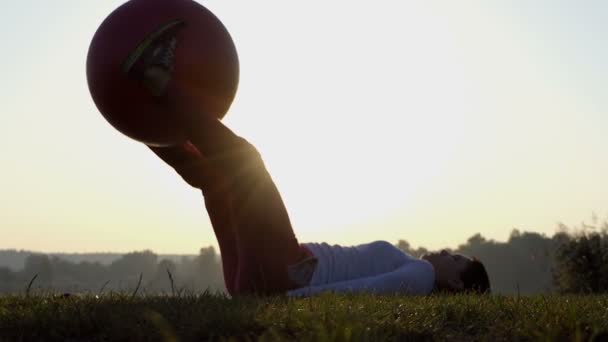 Uma mulher esbelta levanta seu fitball com as pernas no por do sol em slo-mo — Vídeo de Stock