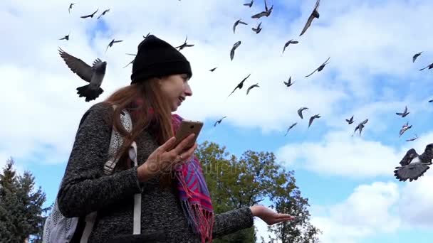 Una mujer feliz con un smartphone se ríe mientras alimenta a las palomas en slo-mo — Vídeo de stock