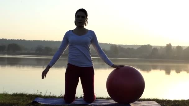 Schlanke Frau kniet und beugt sich bei Sonnenuntergang neben einem Fitball zur Seite — Stockvideo