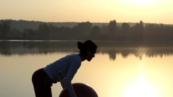Slender woman raises her fitball up at sunset — Stock Video