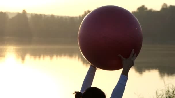 Mulher esbelta levanta seu fitball enquanto deitado ao pôr do sol em slo-mo — Vídeo de Stock