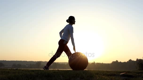 Mujer delgada se para, se arrodilla, rueda su fitball al atardecer en slo-mo — Vídeo de stock