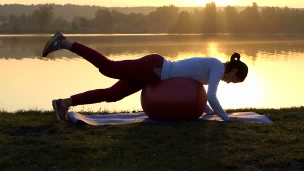 Sportive žena leží na zdravotním balonu a vyvolává nohu při západu slunce — Stock video