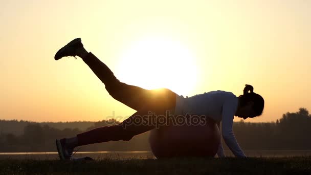 Slanke vrouw ligt op een fitball en werpt een been bij zonsondergang — Stockvideo