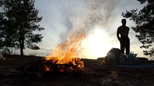 Uma fogueira com garfos brilhantes de chama e um homem ao pôr-do-sol em câmara lenta . — Vídeo de Stock