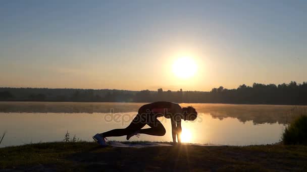 Uma mulher magra treina fitness em um banco de lago ao pôr do sol — Vídeo de Stock