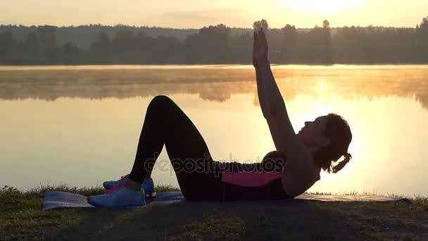 Slim woman lies on a lake bank and raises head at sunset — Stock Video