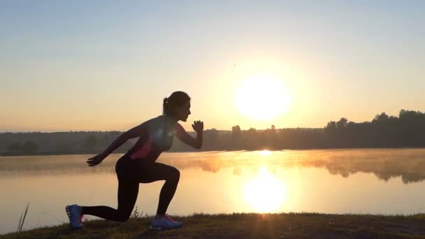 Frau springt bei Sonnenuntergang auf einem Bein am Ufer eines Sees — Stockvideo