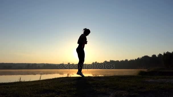 Femme saute sur son tapis sur une rive de lac au coucher du soleil — Video