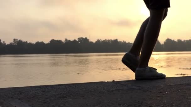 Pernas masculinas caminham e dançam em uma margem do rio ao pôr do sol no verão em Slo-Mo — Vídeo de Stock