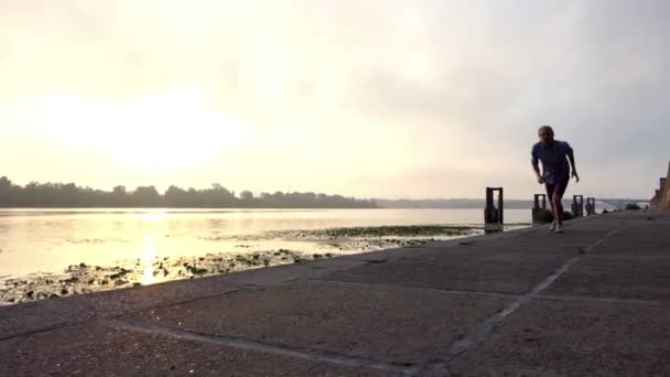 Valiente deportista corre en la orilla del río con losas de hormigón al atardecer en Slo-Mo — Vídeo de stock