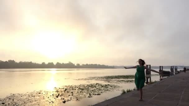 Mulher grávida feliz corre na margem do rio em um vestido verde ao pôr do sol em Slo-Mo — Vídeo de Stock