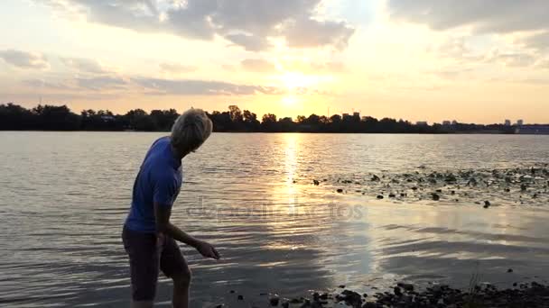 Young Man está parado en una orilla del río al atardecer. arroja una piedra en Slo-Mo — Vídeo de stock